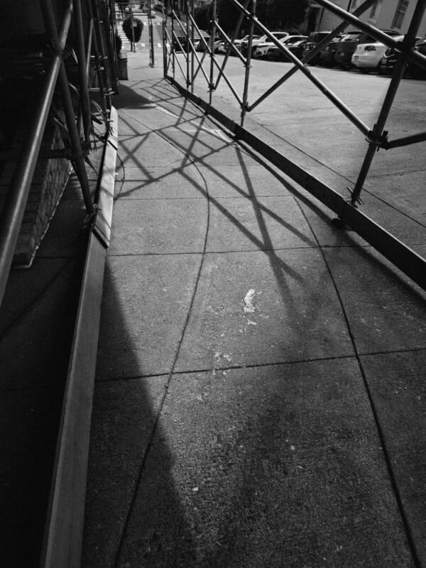 Black and white photo of a sidewalk partially covered by metal scaffolding, casting geometric shadows on the pavement. Cars are parked on the street in the background, and a few people walk in the distance.