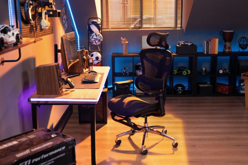A modern gaming setup features a mesh ergonomic chair at a wooden desk with a computer, keyboard, and speakers. LED lights, shelves with books and decor, and gaming accessories fill the cozy, organized room.