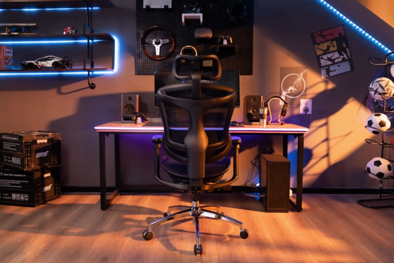 A modern gaming setup features a black ergonomic chair at a desk with dual speakers, a monitor, and LED lighting. Shelves with model cars, sports balls, and gaming accessories decorate the room.