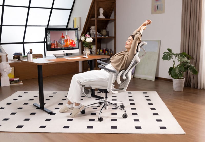 A person in a home office stretches back in an ergonomic chair at a desk with a computer. The room has wooden floors, a patterned rug, plants, and shelves with art supplies near a large window.
