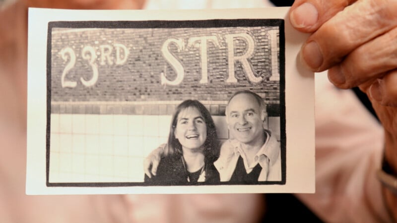 Una mano sostiene una fotografía en blanco y negro de una mujer sonriente y un hombre posando frente a una pared. "RUE N° 23" visible, pista "Calle 23.