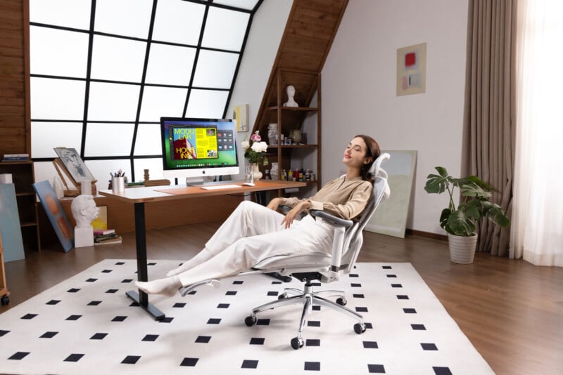 A woman reclines in an ergonomic office chair with her eyes closed, relaxing in a modern home office with a computer on a wooden desk, large window, plants, and decorative items around her.
