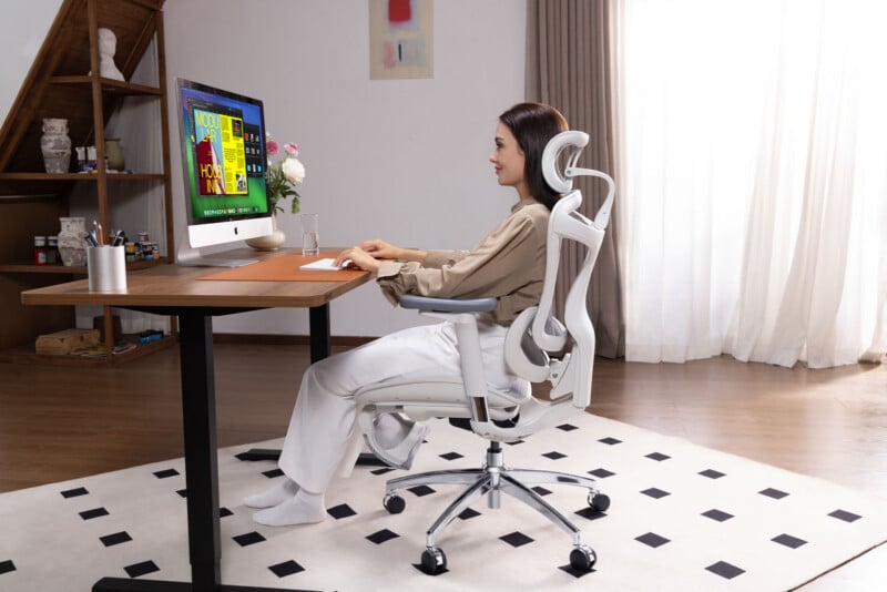 A woman sits in an ergonomic office chair at a desk, working on a desktop computer in a bright, modern room with wooden floors, a large window with curtains, and a black-and-white rug.