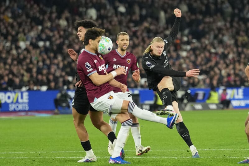 A soccer player in black kicks the ball while players in maroon and white jerseys attempt to block during a match, with a crowded stadium in the background.