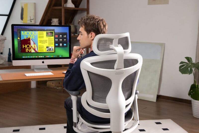 A person sits in a modern ergonomic office chair at a wooden desk, looking at a large computer monitor displaying a colorful design project in a bright, stylish room.