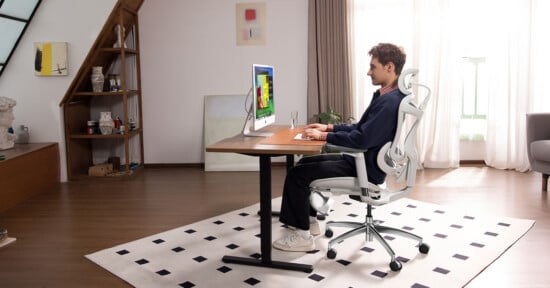 A person sits at a desk working on a computer in a modern, bright home office with wooden floors, a patterned rug, and shelves under a staircase. Natural light enters through large windows with curtains.