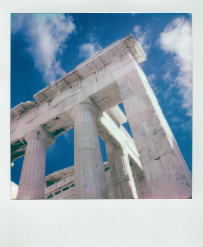 Fotografía estilo Polaroid tomada desde un ángulo bajo que muestra las antiguas columnas de mármol y las ruinas del Partenón bajo un cielo azul vibrante con nubes dispersas.