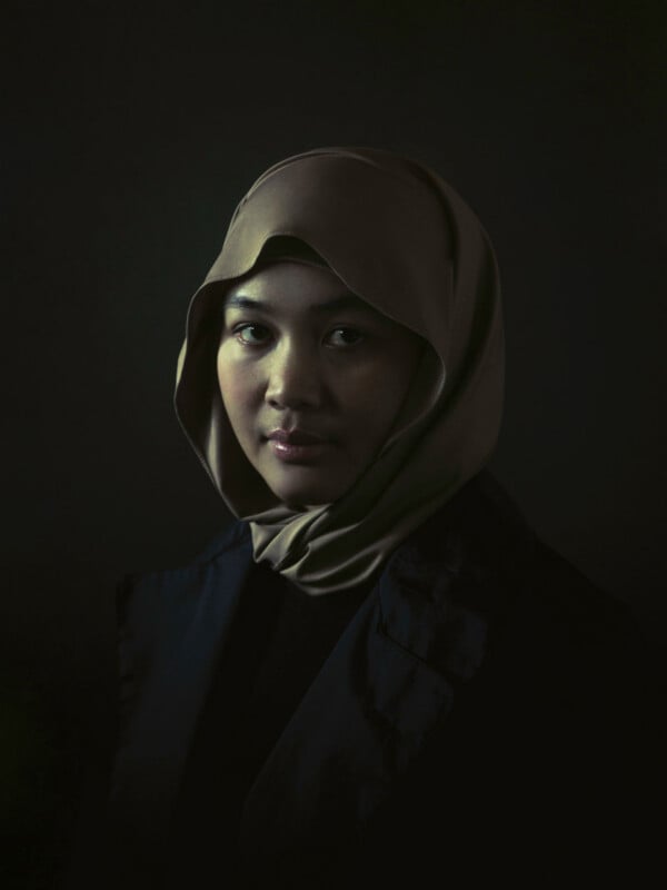 A woman wearing a taupe hijab and dark clothing gazes softly at the camera against a dark, shadowy background. Soft lighting highlights her serene expression.