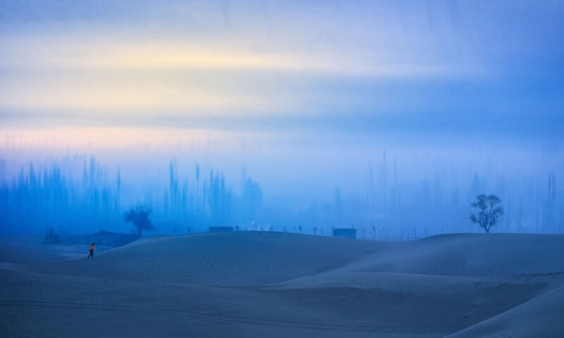 A person walks alone on rolling sand dunes under a blue misty sky, with scattered trees and small buildings in the distance, creating a tranquil and dreamlike landscape.