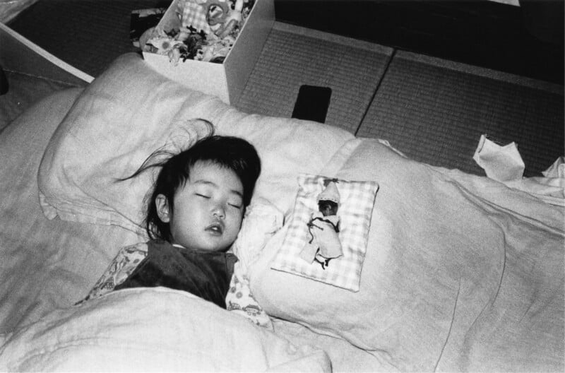 A young child sleeps peacefully on a bed with a patterned pillow featuring a doll design beside them. A box with various items sits nearby on a tatami mat floor. The scene is in black and white.