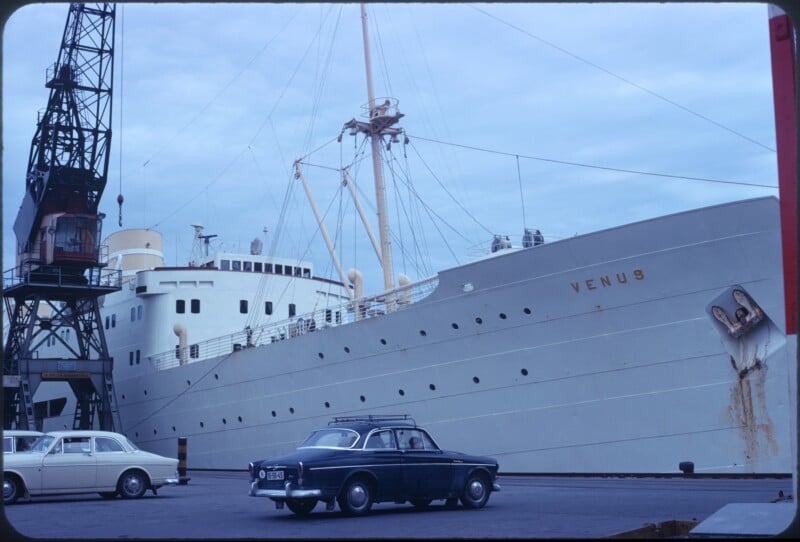A large white ship named "VENUS" docked at a port, with two classic cars parked in the foreground and a crane visible on the left side. The sky is overcast.