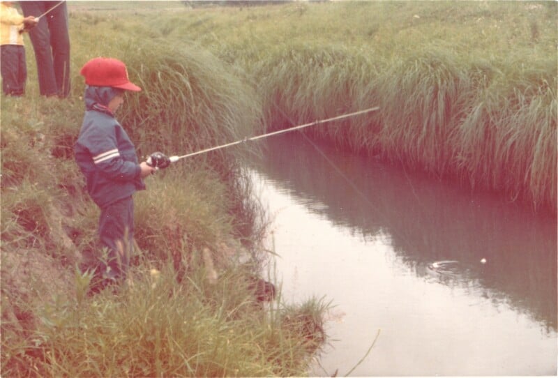 A young child wearing a red hat and blue jacket stands on a grassy bank, fishing in a narrow stream. The fishing line stretches out over the water, with ripples visible where it lands.
