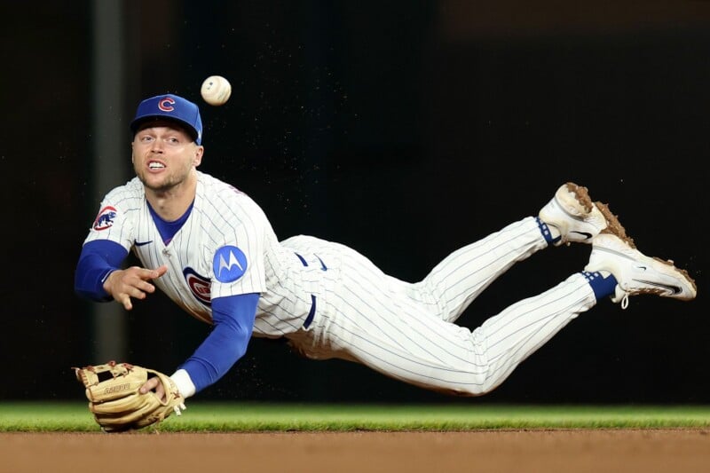 A Chicago Cubs baseball player in a white pinstripe uniform dives in midair, reaching with his glove toward a baseball as it flies past, during a game.
