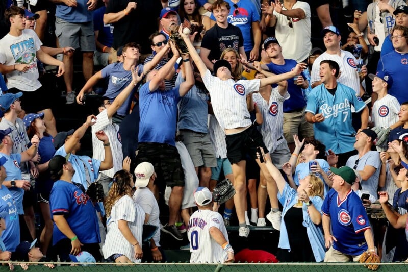 A crowd of excited baseball fans in Cubs and Royals jerseys reach up to catch a ball in the stands, with arms outstretched and faces full of excitement and anticipation.