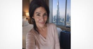 A woman with dark hair and a light pink top smiles for a selfie in a hotel room with a view of the Burj Khalifa and cityscape through a large window. A bed and suitcase are visible in the background.