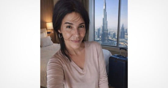 A woman with dark hair and a light pink top smiles for a selfie in a hotel room with a view of the Burj Khalifa and cityscape through a large window. A bed and suitcase are visible in the background.