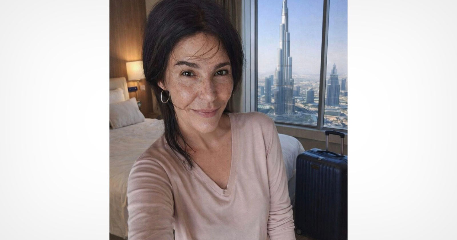 A woman with dark hair and a light pink top smiles for a selfie in a hotel room with a view of the Burj Khalifa and cityscape through a large window. A bed and suitcase are visible in the background.