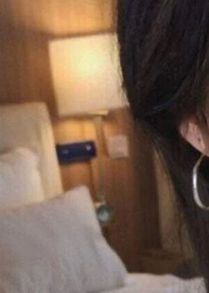Close-up of a person’s ear with a large hoop earring; background shows a bed with white pillows and sheets, a bedside lamp, and a wooden headboard. Only part of the person’s face is visible.