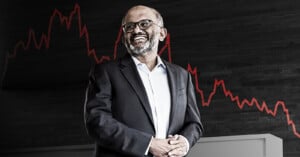 A smiling man in a suit and glasses stands in front of a dark background with a red fluctuating line graph, suggesting changes in stock or financial performance.