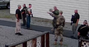 A group of armed law enforcement officers stand and talk outside near a white metal building on a gravel path, with some facing each other and one officer pointing.