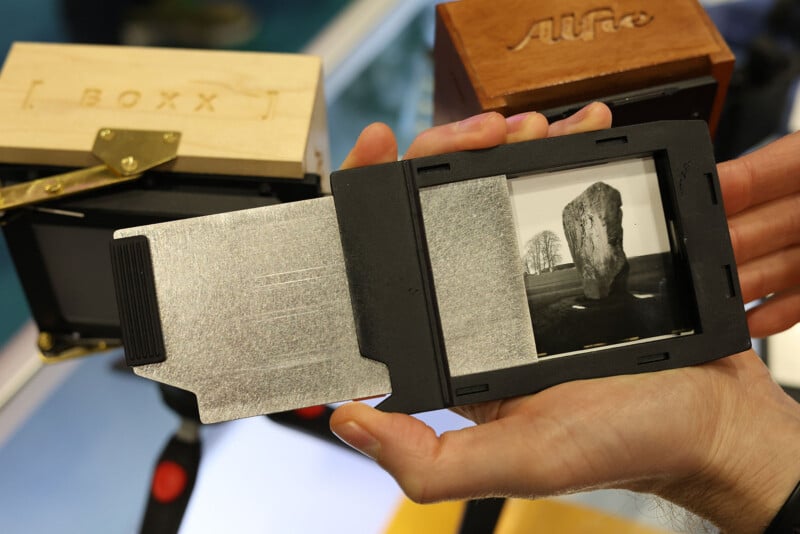 A hand holds a photographic plate holder with a black and white image of a large standing stone and trees; wooden camera equipment is visible in the background.