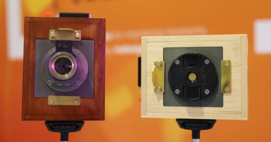 Two vintage-style box cameras with wooden bodies—one in dark wood, one in light wood—are mounted on tripods against an orange, blurred background.