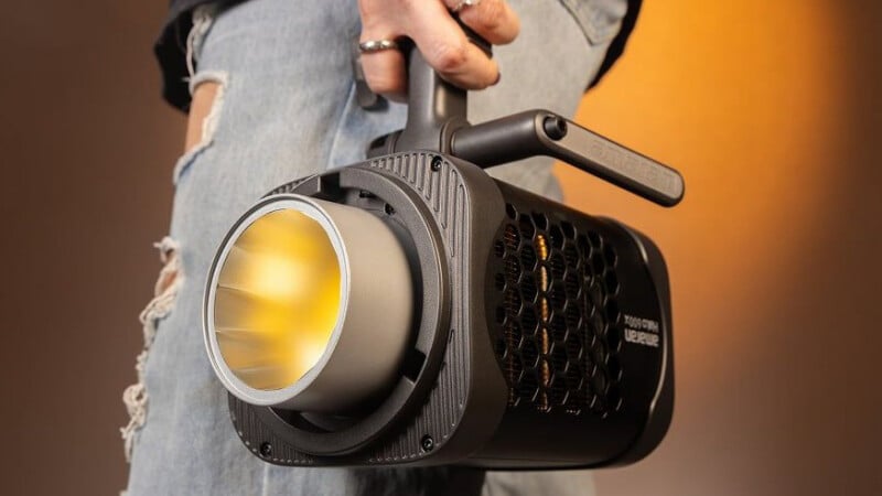 A person wearing ripped jeans holds a black, cylindrical LED studio light with a handle, pointed downward. The background has a warm, orange gradient.