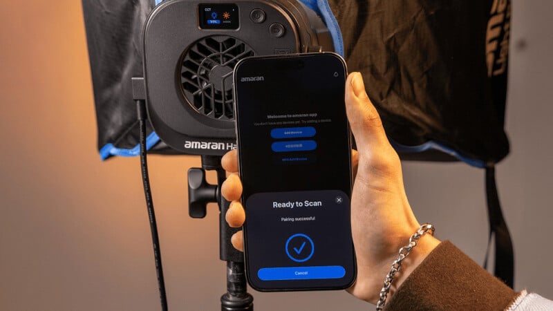 A person holds a smartphone displaying a pairing screen in front of an Aputure Amaran LED studio light, preparing to connect the phone to the light via a mobile app.