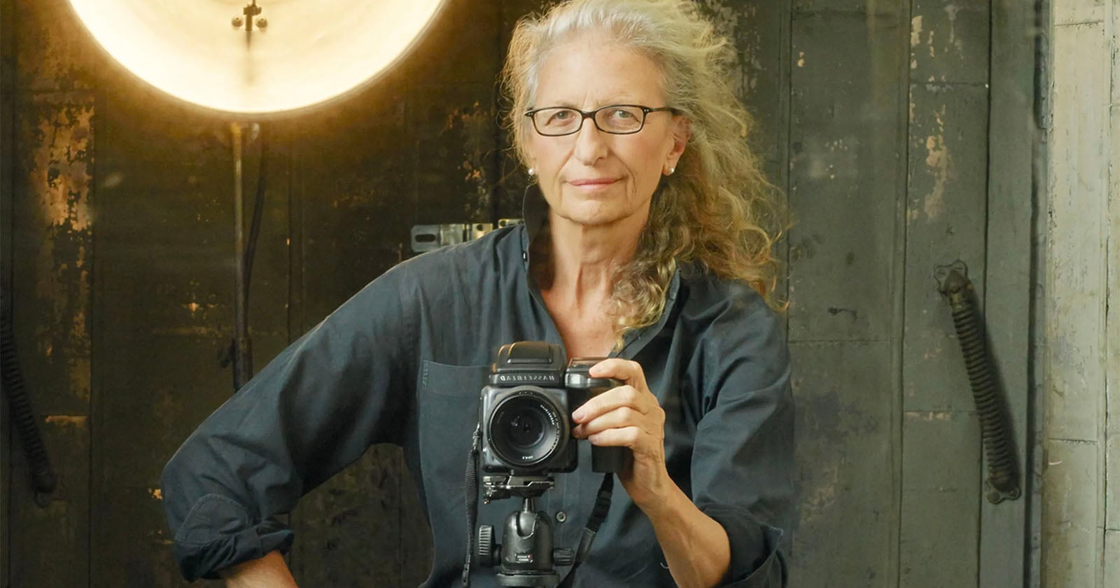 A woman with curly gray hair and glasses stands in front of a camera on a tripod, posing for a self-portrait. She wears a dark shirt, with a round light behind her and a rustic, industrial background.