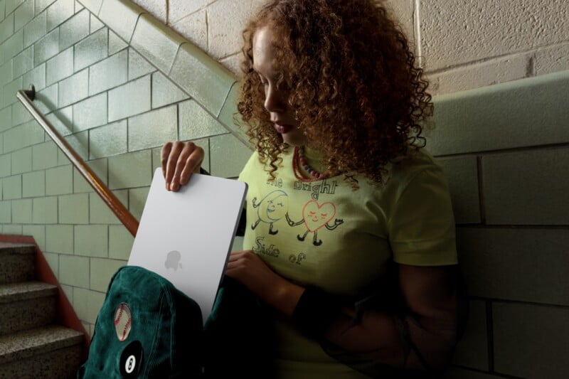 A young person with curly hair stands on a stairwell, pulling a silver laptop out of a green backpack. They are wearing a light green shirt with cartoon faces and text, against a background of green and beige tiled walls.