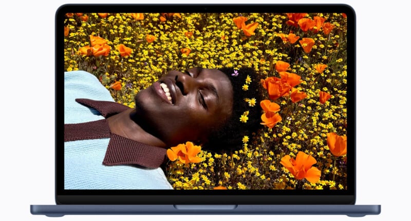 A smiling person lies in a field of orange and yellow flowers, shown on the screen of a laptop.