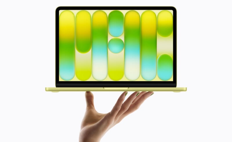 A hand holds up a slim, yellow laptop with an open screen displaying an abstract design of green, yellow, and blue vertical ovals and gradients. The background is plain white.