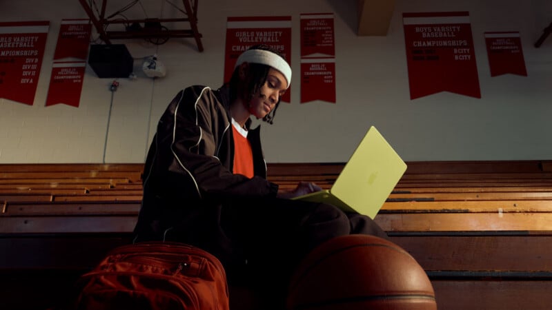 A person in athletic wear sits on bleachers with a basketball and orange backpack, typing on a yellow laptop. Red sports championship banners hang on the wall behind them in a gymnasium.