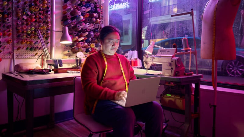 A person wearing a red sweater and yellow measuring tape works on a laptop in a sewing studio, surrounded by thread spools, a sewing machine, and a dress form, with purple lighting and rain outside the window.