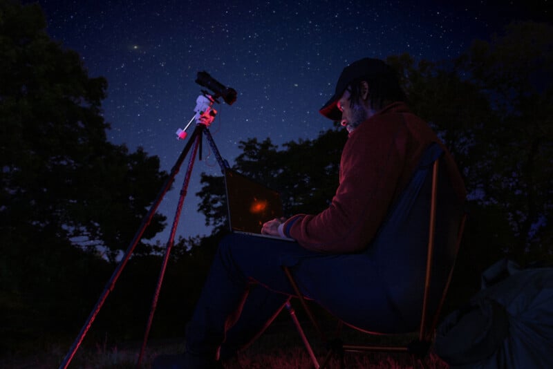 A person sits outdoors at night using a laptop, illuminated by red light, next to a telescope on a tripod, with a star-filled sky in the background and trees silhouetted in the dark.