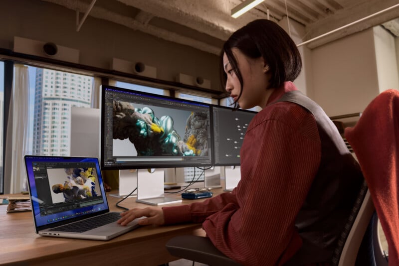 A person sits at a desk in an office, using a laptop and two large monitors to create 3D digital art. The screens display a colorful, detailed 3D model, with city buildings visible through the windows behind them.