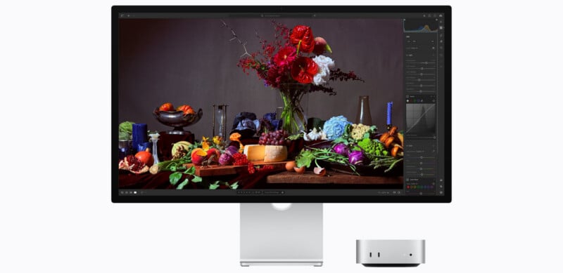A computer monitor displays a vibrant still-life image of flowers, fruits, and vegetables. A small silver desktop computer sits below the screen against a plain white background.