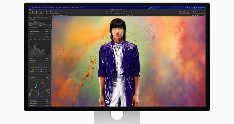 A computer monitor displays a vivid portrait of a person in a shiny purple jacket, standing against a vibrant, colorful background with orange and green clouds; photo editing software interface is visible on the screen.