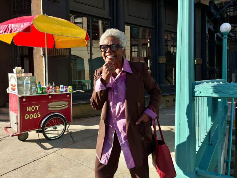An older woman with gray hair, wearing sunglasses, a brown suit, and purple shirt, smiles and stands near a red hot dog cart with a yellow-and-red umbrella on a sunny city sidewalk.