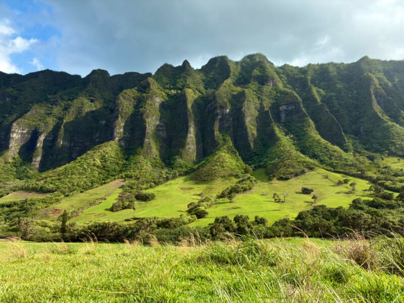 Towering green cliffs with sharp ridges rise above a grassy field under a cloudy sky, with sunlight casting dramatic shadows on the mountainside.