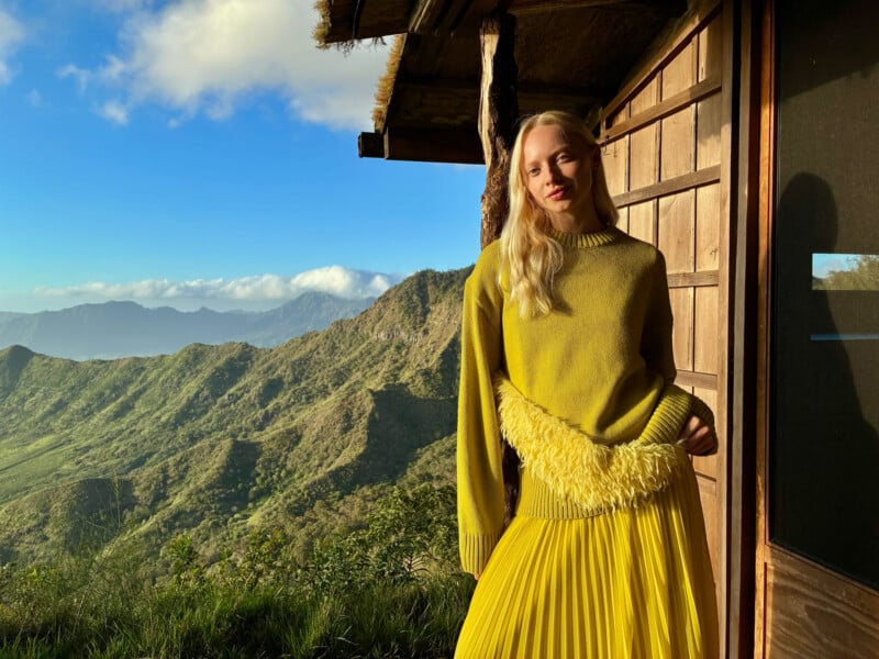 A woman in a yellow sweater and skirt stands by a wooden building, bathed in sunlight, with green mountains and a blue sky with clouds in the background.