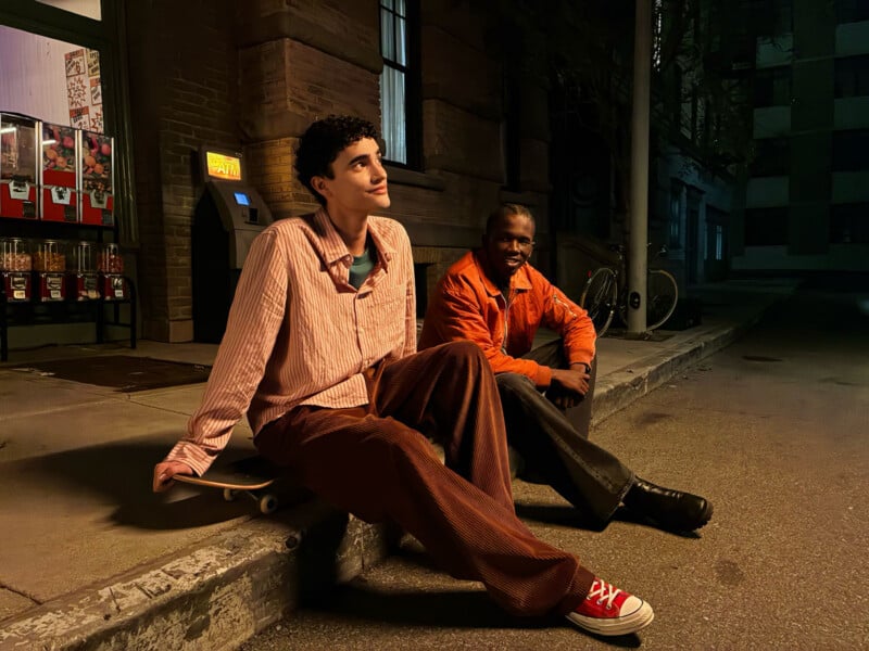 Two young men sit on a curb at night under streetlights; one is sitting on a skateboard and smiling, while the other, in an orange jacket, sits slightly behind him. They appear relaxed and are near a vending machine.
