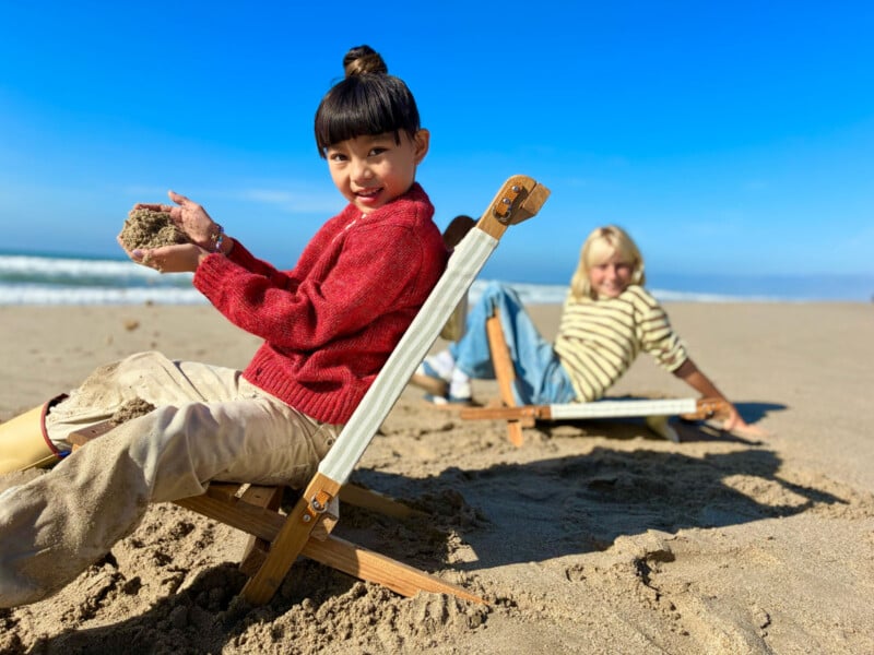 Two children sit on wooden beach chairs on sandy beach under clear blue sky; one child in a red sweater holds sand and smiles at the camera, while the other sits in the background, slightly out of focus.