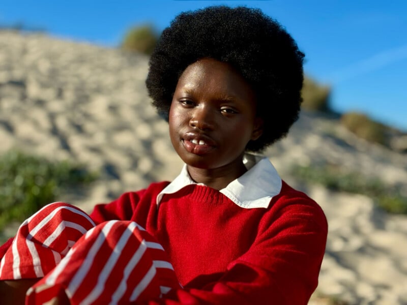 A person with a natural afro hairstyle wearing a red sweater with a white collar and red-and-white striped pants sits outdoors on a sandy landscape under a clear blue sky.