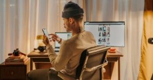 A person wearing a beanie sits at a desk, using a smartphone. Two monitors, a laptop, camera, and headphones are on the desk. The room is warmly lit with curtains in the background.