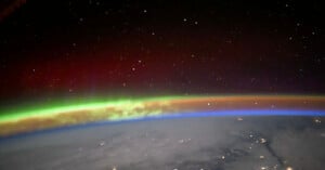 View of Earth from space showing a colorful aurora with green, red, and orange hues above the planet’s curved horizon, set against a star-filled black sky. Some city lights are visible on the ground below.