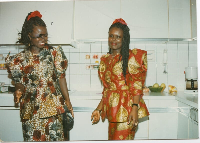 Two women stand in a white tiled kitchen, both wearing vibrant, patterned dresses with puffed sleeves. One woman smiles while holding a black purse; the other stands with hands relaxed, wearing a matching headpiece.
