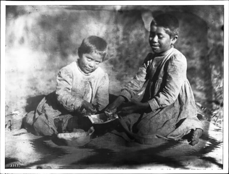 Two young children sit on a patterned blanket, sharing food from a bowl. Both wear simple, worn clothing and appear focused on the meal, with a soft, blurred background behind them.