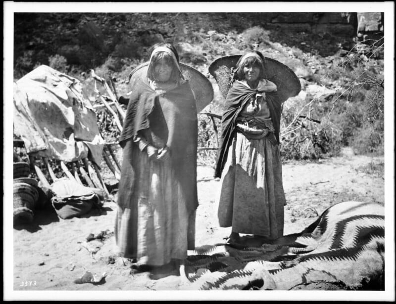 Two Indigenous women stand outdoors wearing long dresses and shawls, each with a large woven basket on their backs. Behind them are makeshift wooden structures and a patterned blanket on the ground.