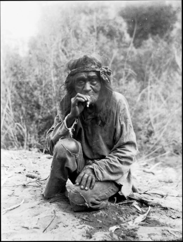 An elderly man with long hair and a headband squats outdoors, dressed in worn clothes. He looks directly at the camera while touching his mouth, surrounded by dense foliage. The photo is in black and white.